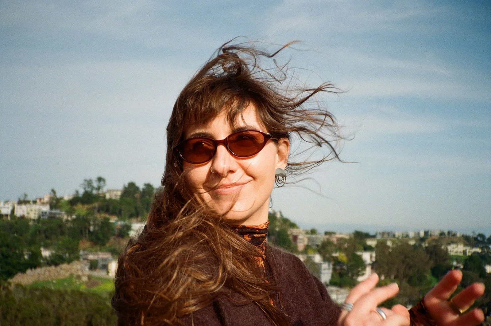 A woman with long curly hair wearing sunglasses smiling outdoors, with a hilly landscape and buildings in the background.