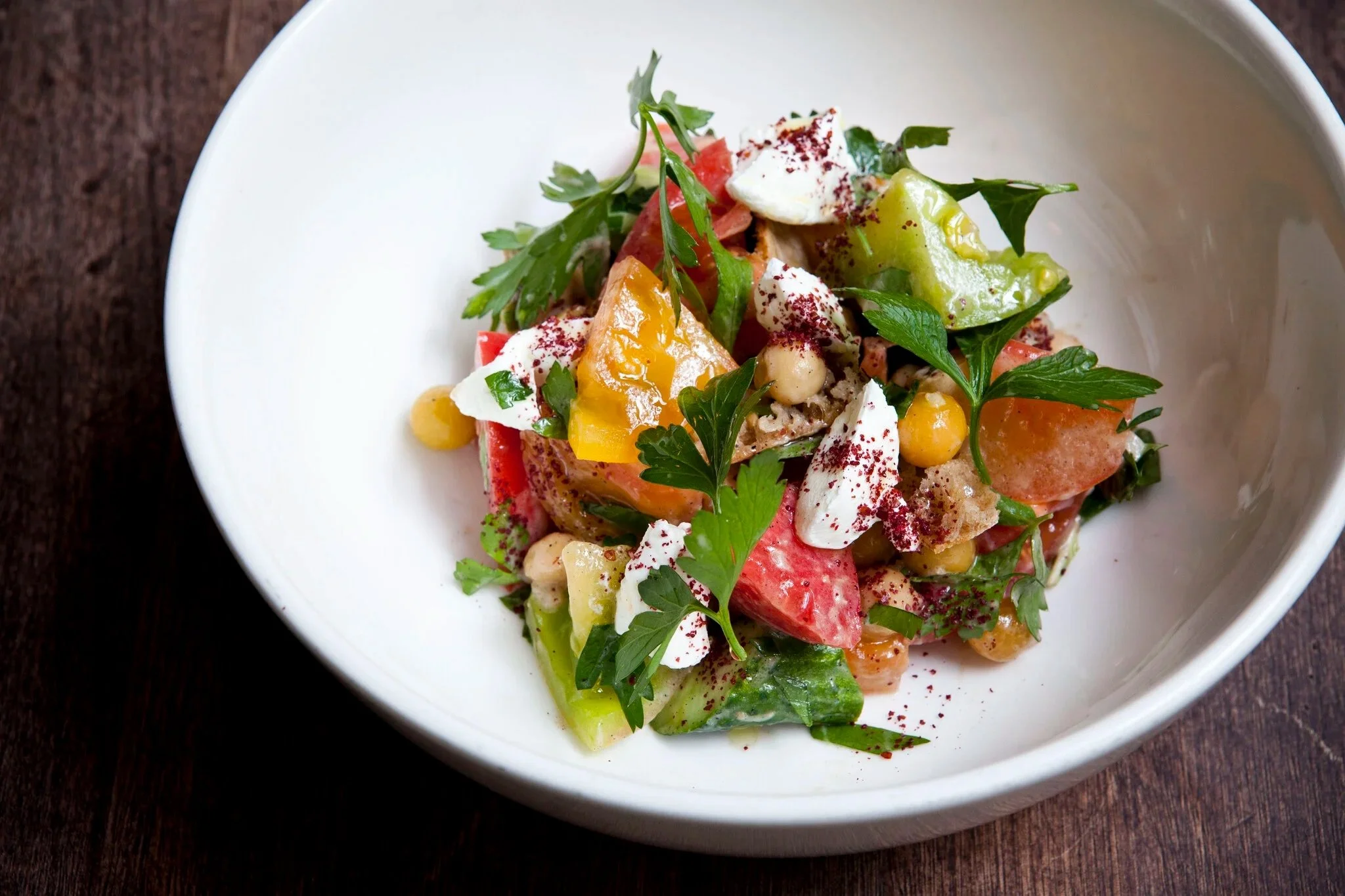 Ein Salat mit Tomaten, Gurken, weißen Bohnen, grüner Sauce, frischen Kräutern und geräuchertem Fisch in einer weißen Schüssel auf einem Holztisch.