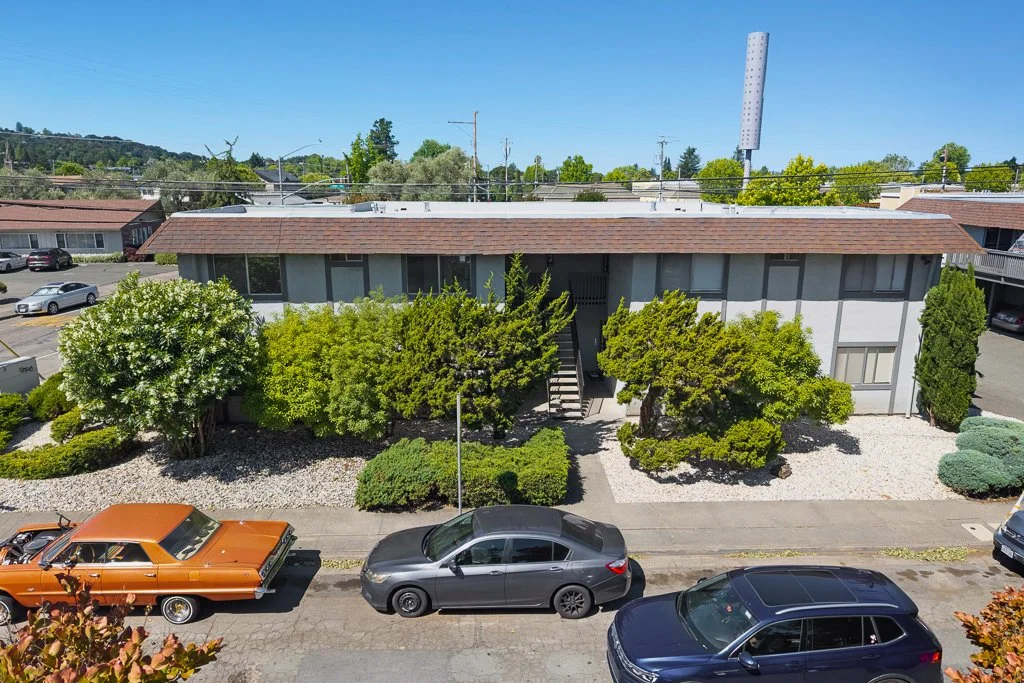 A two-story apartment building with a red-tiled roof, surrounded by bushes and trees, with a parking lot in front containing several cars, in a suburban area with greenery and clear blue sky.