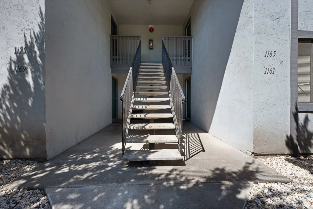 Exterior of an apartment building with stairs leading to a second-floor entrance, two unit numbers 1165 and 1161 on the wall, and shadows from nearby trees.