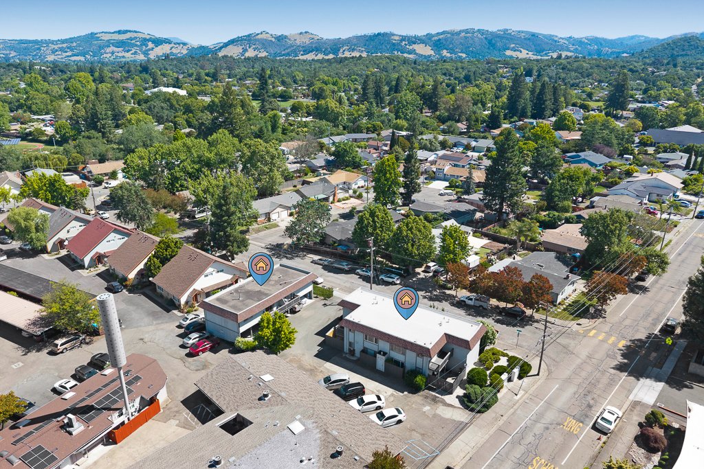 Aerial view of a suburban neighborhood with numerous single-family houses surrounded by trees, with mountains in the background. Two houses are marked with house icons.