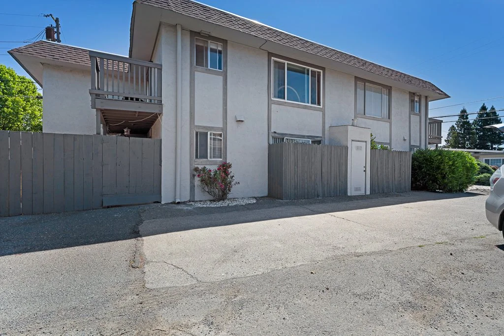 Two-story beige residential building with a sloped roof, balconies, and a fenced yard, on a sunny day.