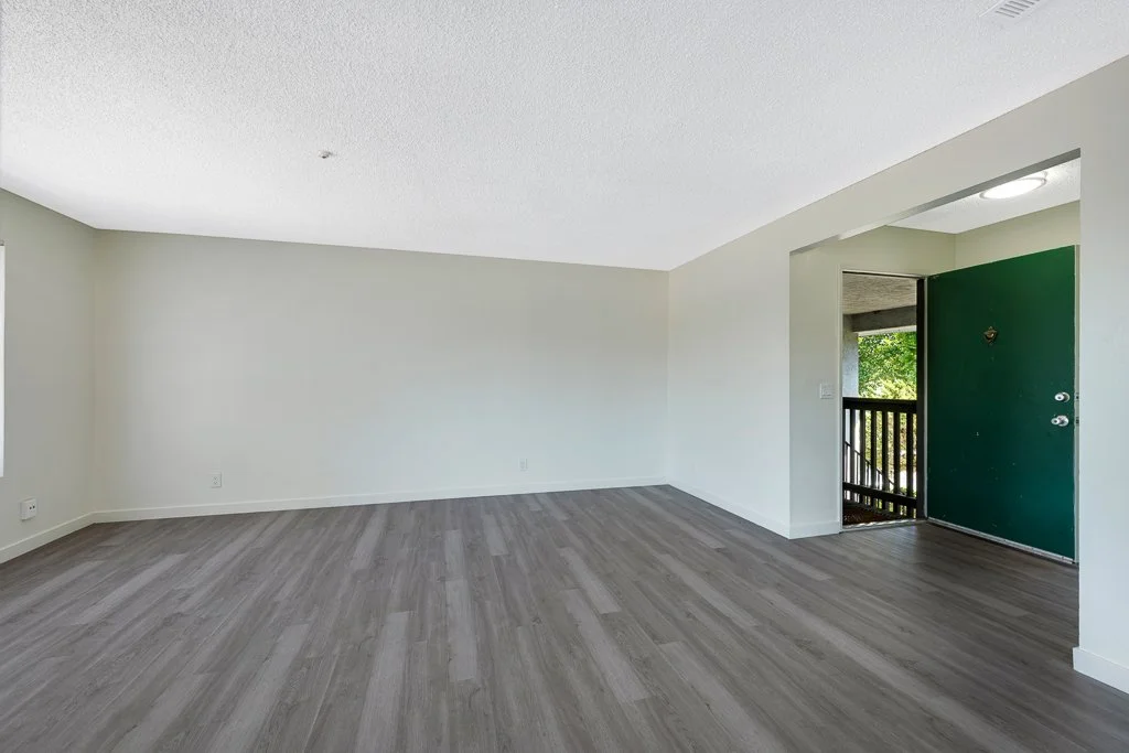 Empty room with gray wooden flooring, white walls, a green front door, and a small balcony visible through the doorway.
