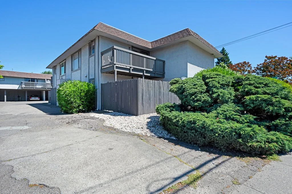 A two-story residential building with a small balcony, surrounded by shrubs and a wooden fence, with parking spaces and a neighboring building in the background.