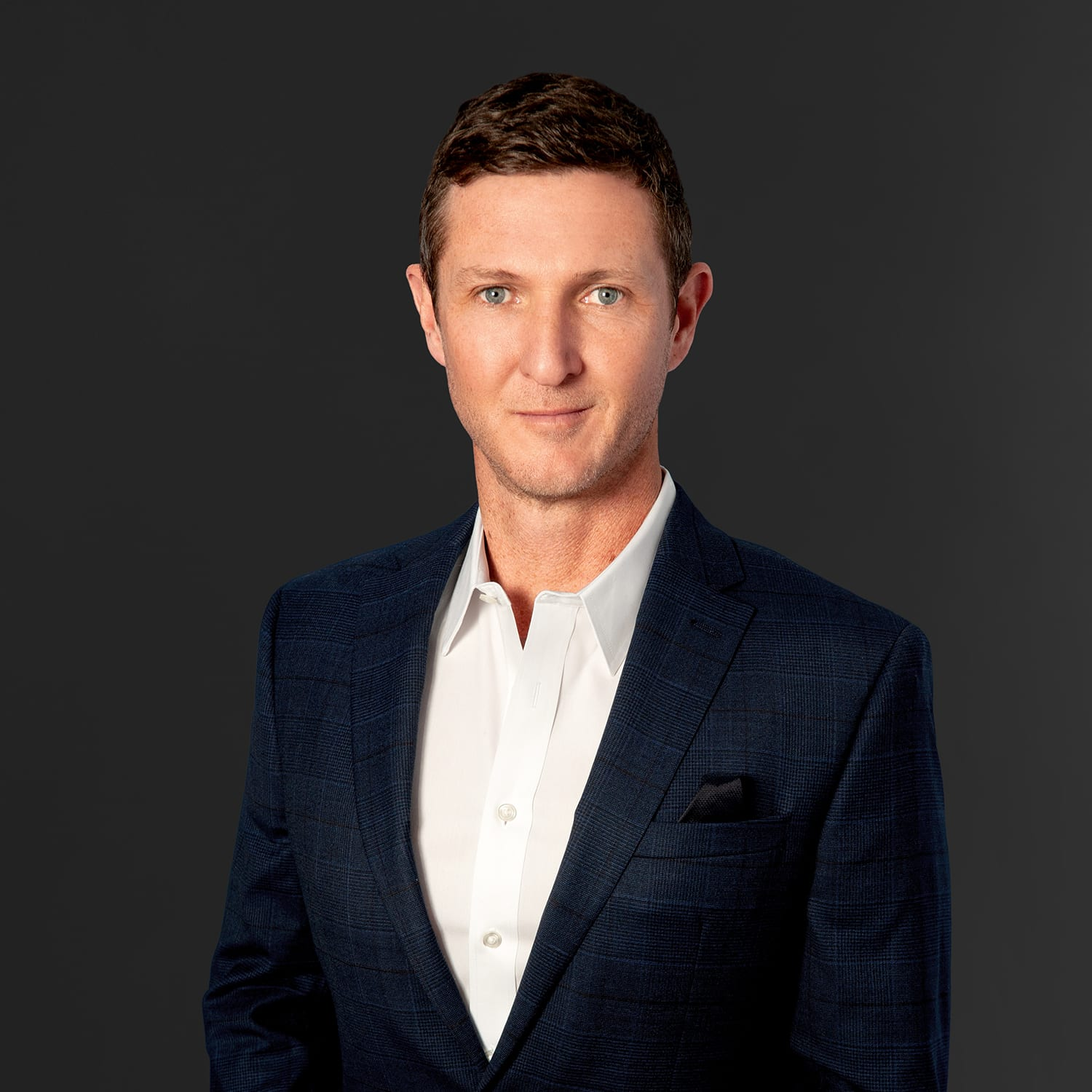 Portrait of a man with short brown hair, blue eyes, wearing a white dress shirt and dark blue checked blazer, against a dark gray background.
