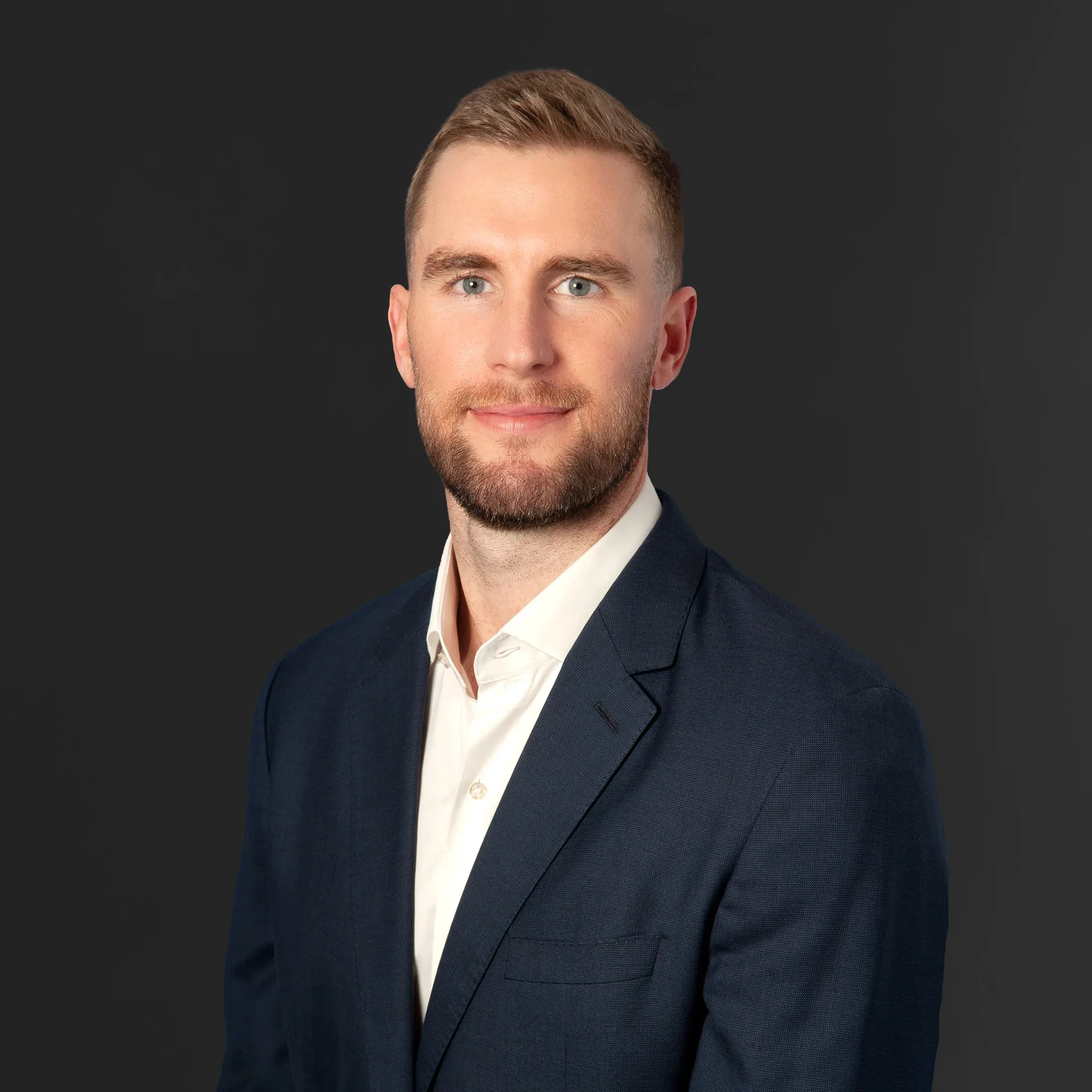 Headshot of a man with light skin, short light brown hair, blue eyes, and a beard, dressed in a white shirt and dark suit jacket, against a dark gray background.