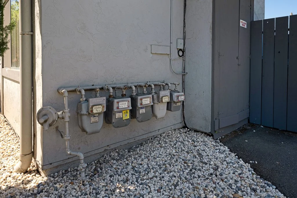 Multiple gas meters mounted on a gray exterior wall with pipes, a large gray utility box, and a gravel ground.