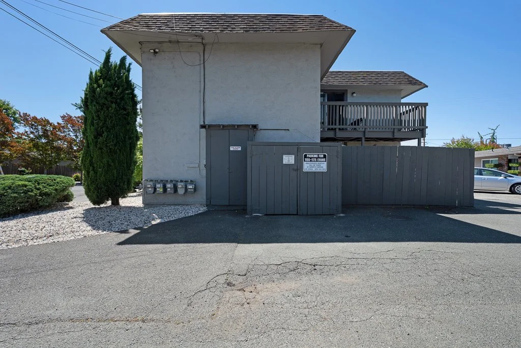 Rear view of a two-story building with an upper balcony, gray exterior, and a fence. There are multiple mailboxes and a sign on the fence indicating parking restrictions. The overall setting appears to be a parking lot or alleyway.