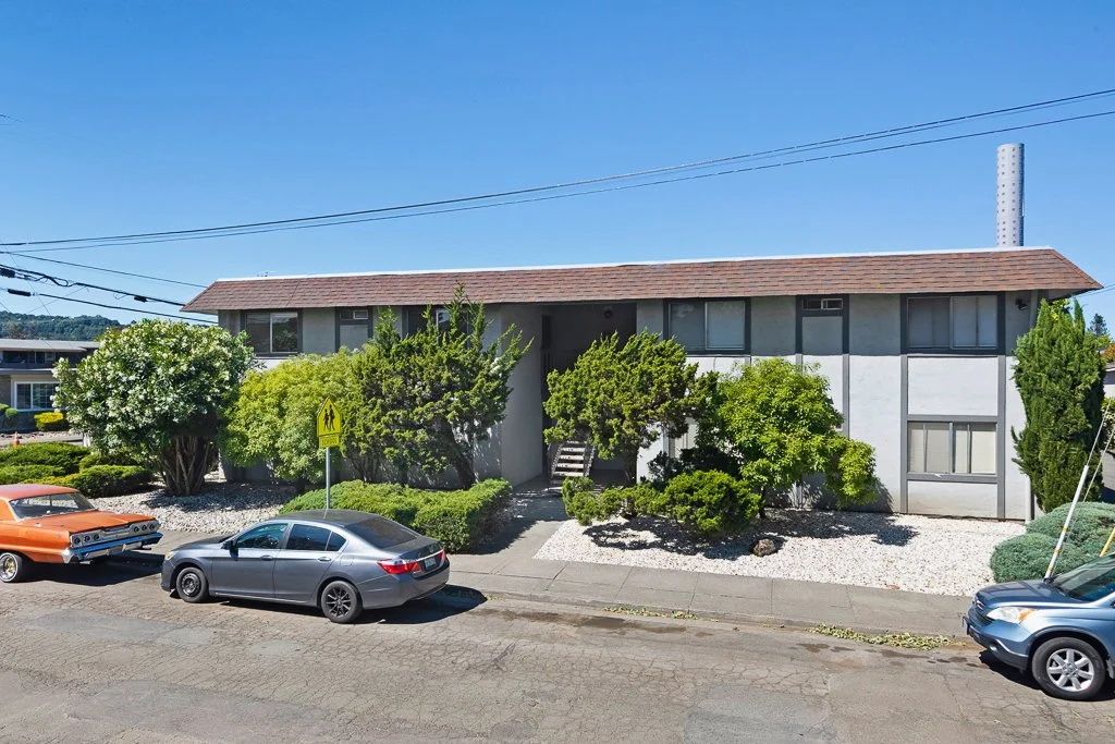 A two-story residential building with a brown roof, surrounded by bushes and trees. There are three cars parked in front: an orange classic car, a gray sedan, and a blue vehicle. There is a yellow pedestrian crossing sign near the bushes and power li