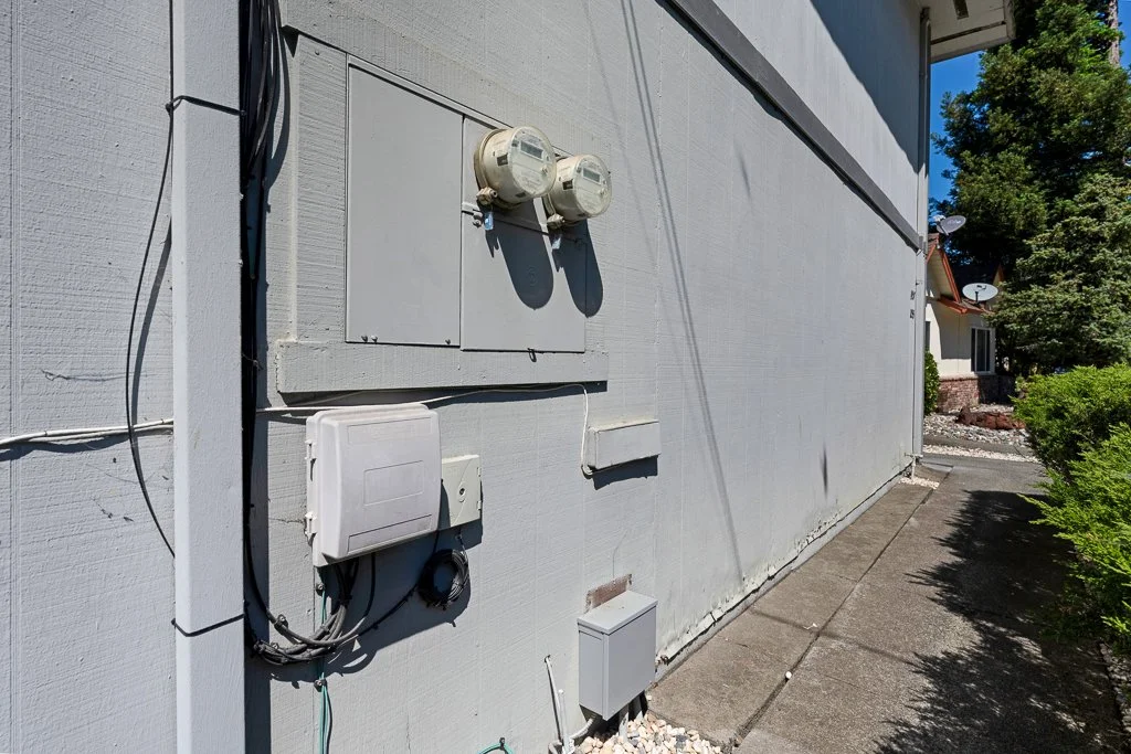 Exterior of a house showing electrical meters, electrical boxes, and wires on the wall next to a sidewalk with bushes and trees in the background.
