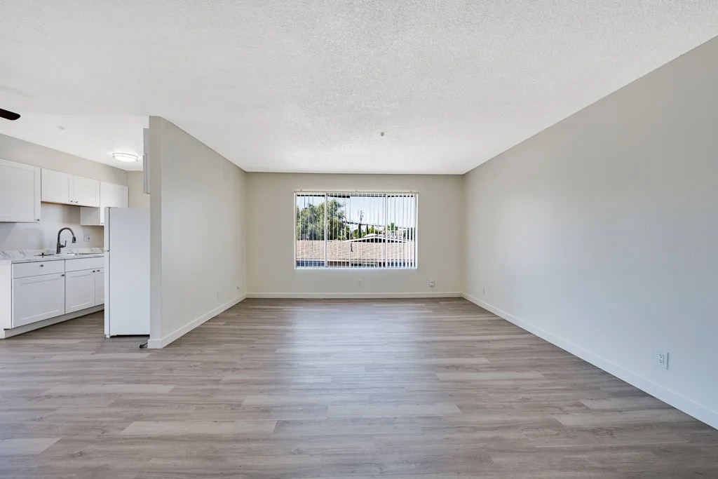 Empty living room with wood flooring, white walls, a large window with vertical blinds, and an open kitchen with white cabinets.