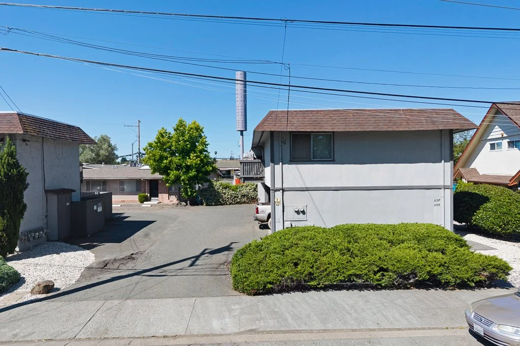 Residential area with two-story gray house, driveway, green bushes, tree, and clear blue sky