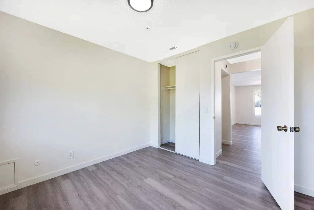 Empty room with white walls, gray wooden flooring, an open closet, and a door leading to a bright room with a window.