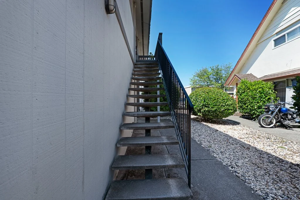 Exterior view of a set of metal stairs with wooden steps attached to the side of a white building, leading to the upper floor, with bushes, gravel, and a motorcycle visible in the background.