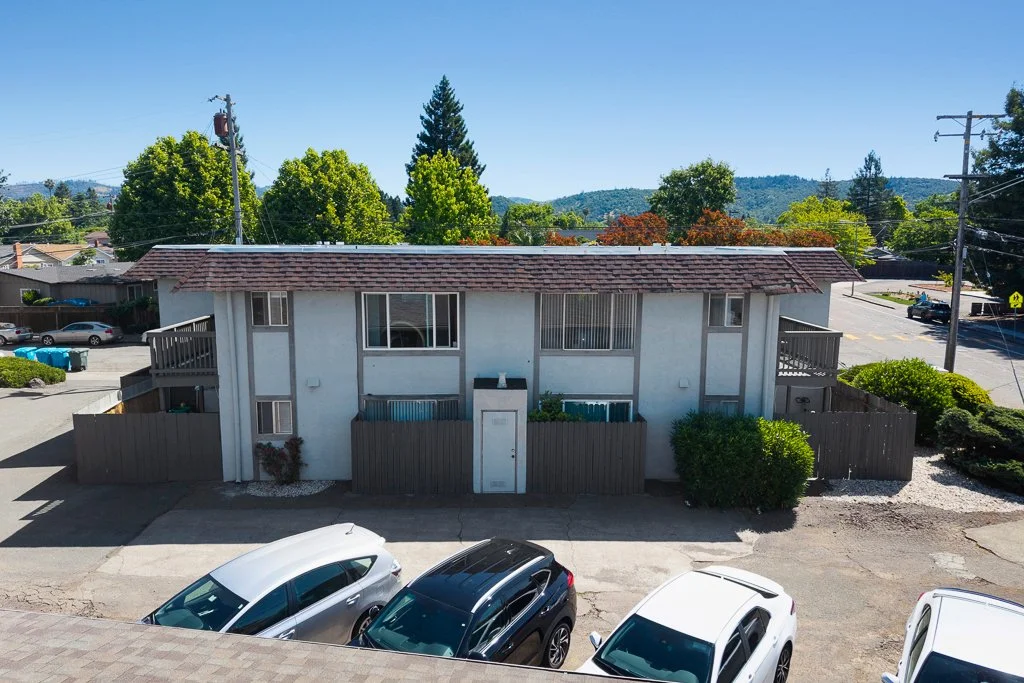 View of a two-story residential apartment building with parking lot and cars in the foreground, trees and hills in the background, under a clear blue sky.