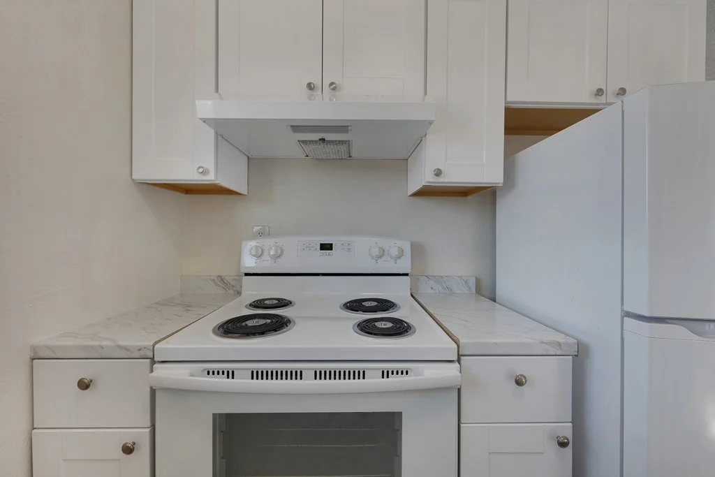White kitchen with a stove, oven, and refrigerator, marble countertops, white cabinets, and a vent hood.