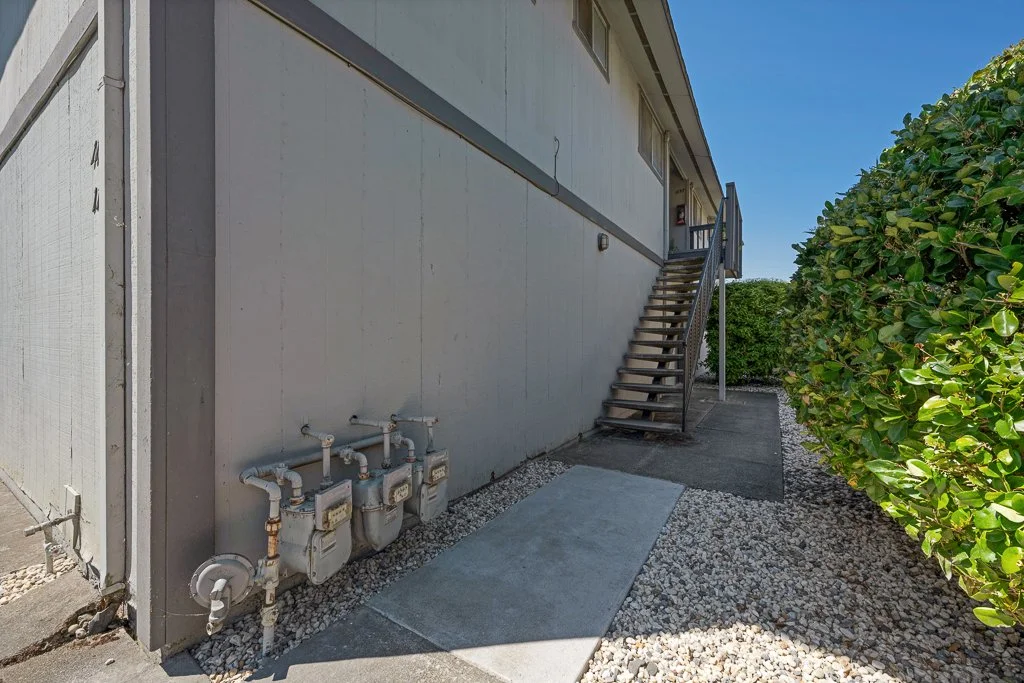Exterior of a building with stairs leading to an upper level, utility meters attached to the wall, and a hedge on the right side.