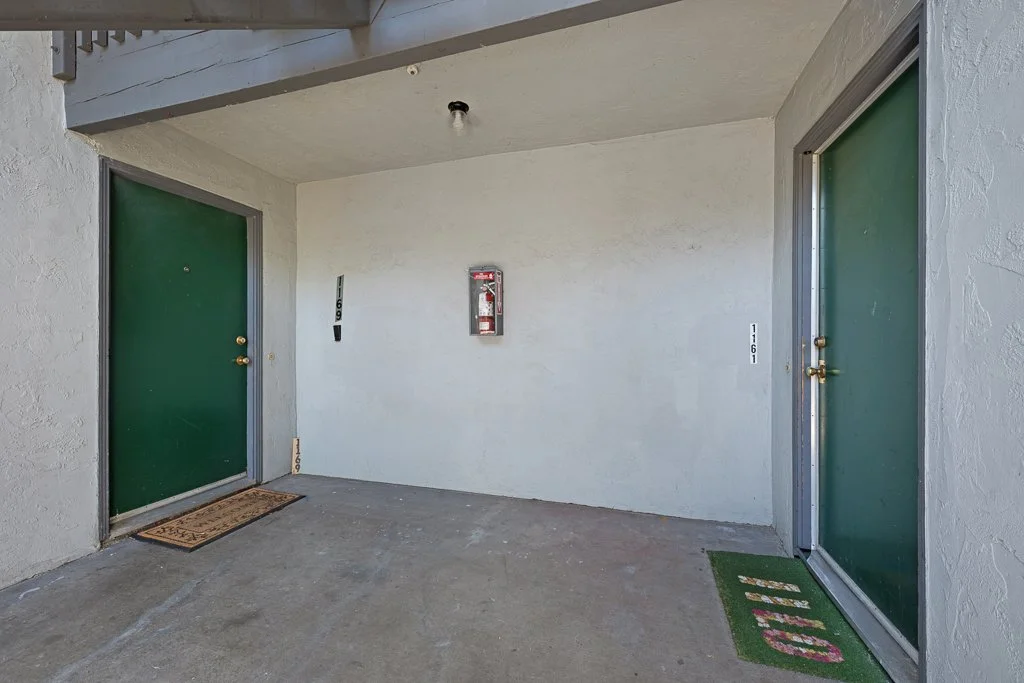 Two green doors with brass doorknobs and two small outdoor mats in front of each door, one with 'WELCOME' and floral decoration, a fire extinguisher in a wall-mounted cabinet, a white textured exterior wall, and concrete ground.