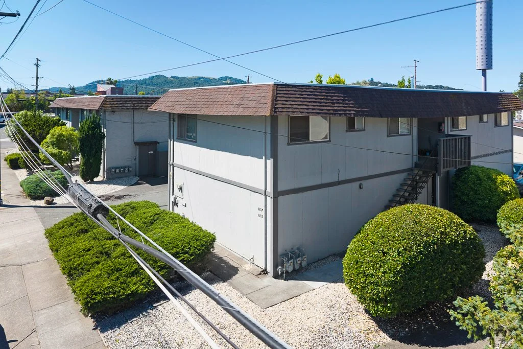 A two-story residential building with a brown shingle roof, white exterior walls, and a wooden staircase leading to a second-floor porch. The property is surrounded by neatly trimmed bushes and small trees, with a sidewalk and power lines in the fore