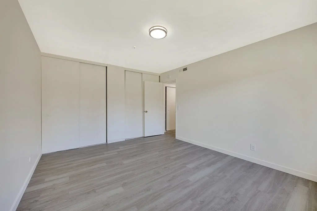 Empty room with light-colored walls, wood flooring, ceiling light fixture, and closet doors, likely a bedroom.