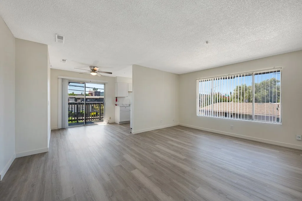 Empty living room with large window and sliding glass door leading to balcony, light-colored laminate flooring, white walls, and ceiling fan.