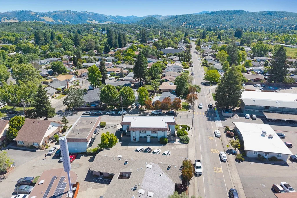 Aerial view of a small town with residential houses, commercial buildings, a main street, and green trees amidst mountain hills in the background.