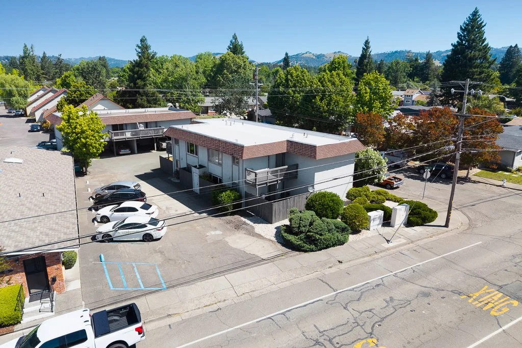 Aerial view of a residential neighborhood with a two-story building, parking lot with several cars, lush green trees, and mountains in the background.