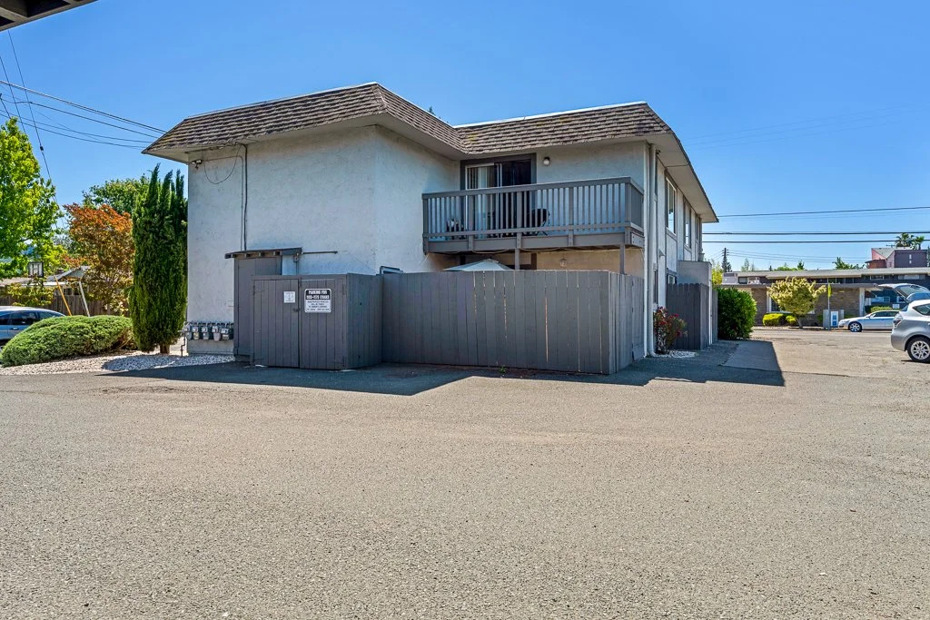 Two-story white residential building with a small balcony, surrounded by a wooden fence, with parking lot and parked cars in the background under a clear blue sky.