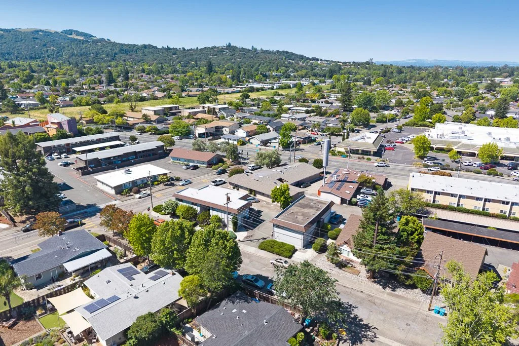 Aerial view of a suburban neighborhood with houses, commercial buildings, trees, and streets, set against a backdrop of hills and clear blue sky.