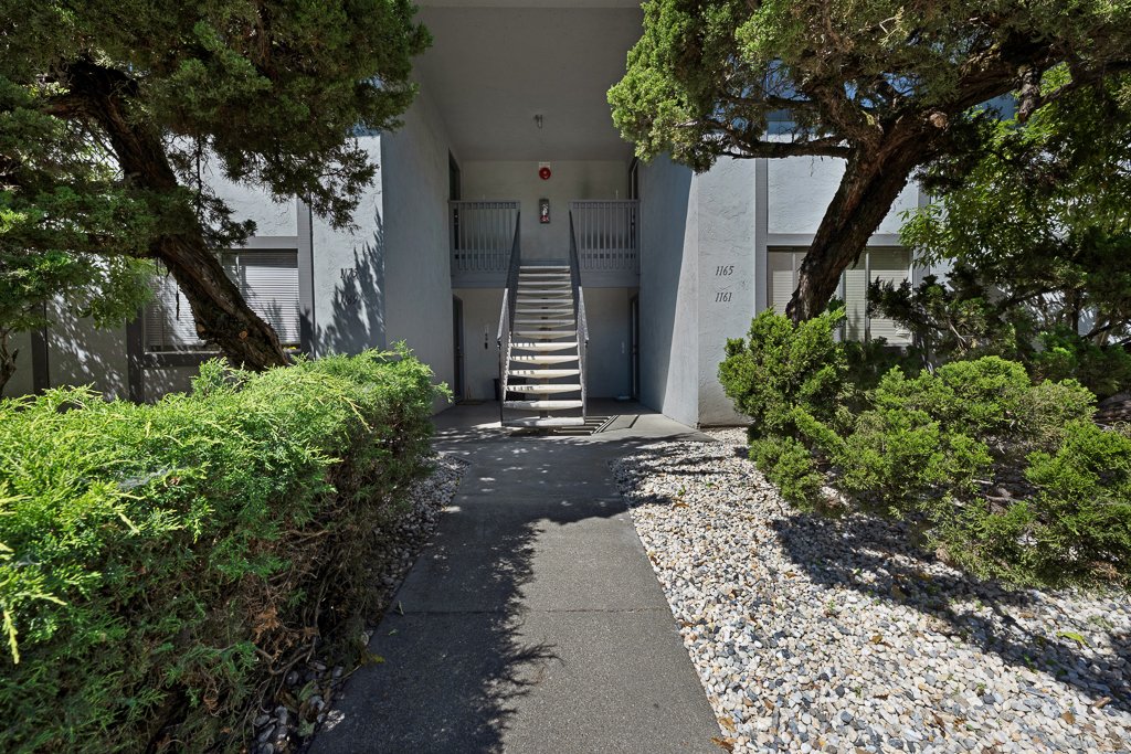 Apartment building entrance with stairs, surrounded by trees and bushes, with two apartment numbers visible.