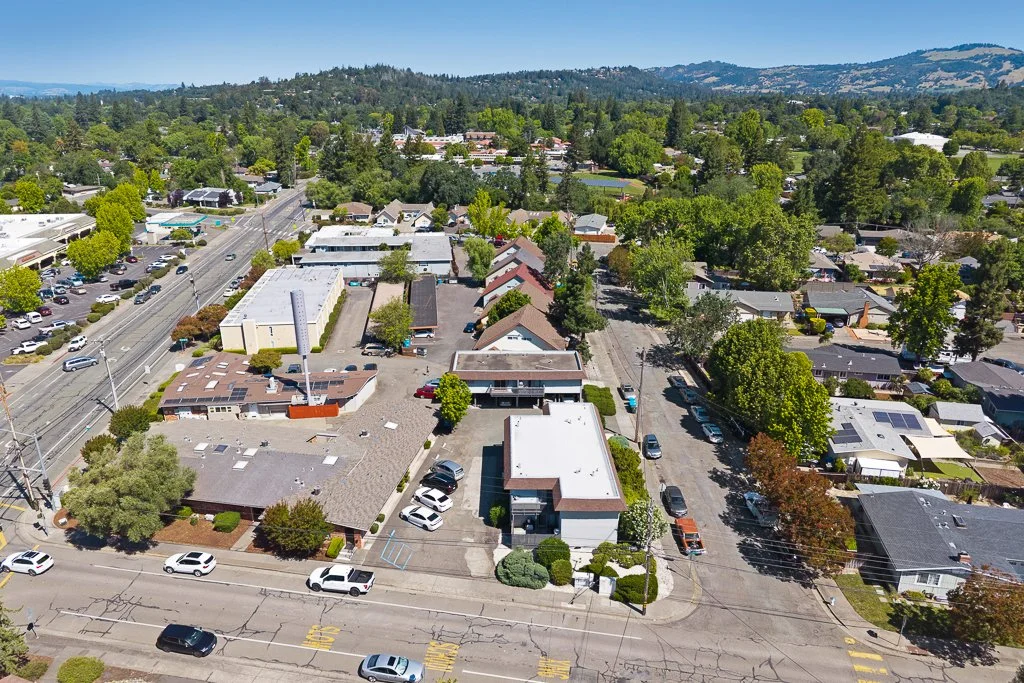 Aerial view of a small town or suburban neighborhood with a mix of commercial buildings, residential houses, and green trees, with hills in the distance.