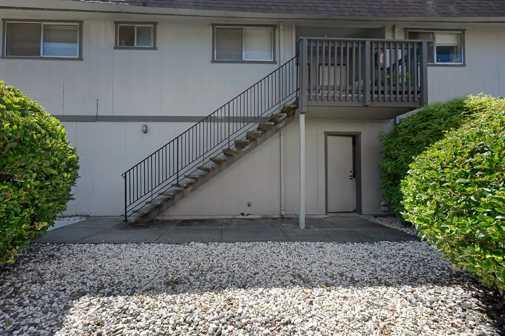 Exterior view of a two-story residential building with stairs leading to the upper balcony, surrounded by bushes and gravel ground.