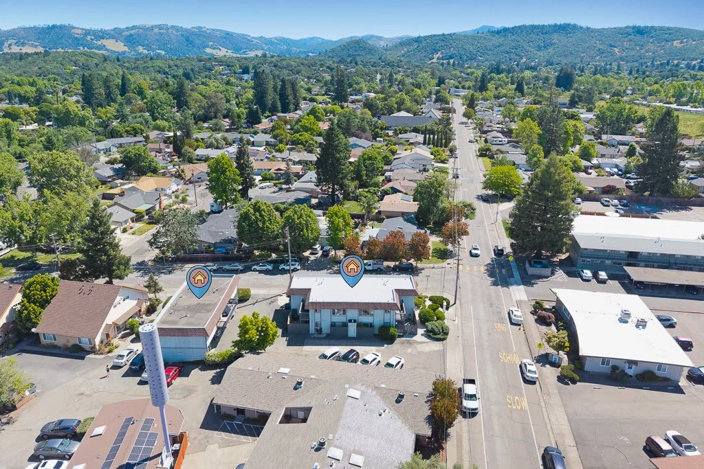 Aerial view of a small town with residential houses, trees, and commercial buildings along a main road with parked cars and some in motion, set against a backdrop of rolling hills and mountains.