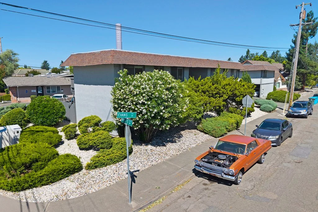 A residential street corner with an orange vintage car parked on the sidewalk, a blue car behind it, and a grey car further down. A house with bushes and trees line the street, with street signs at the corner indicating Evans and Elite streets.