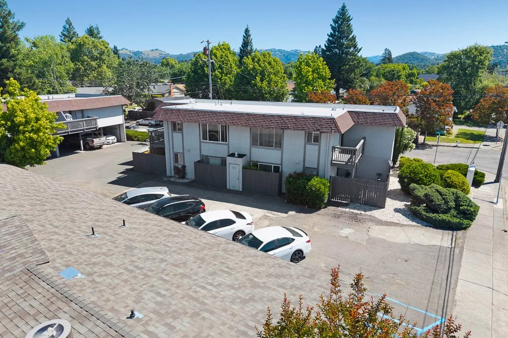 An aerial view of a residential parking lot with several parked cars, apartment buildings, trees, and a clear blue sky in a suburban area.