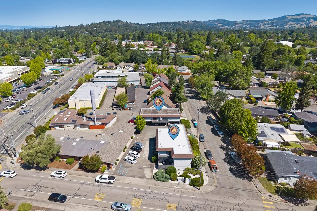 An aerial view of a suburban area with commercial and residential buildings, parking lots, streets, and lush green trees, with hills in the background.