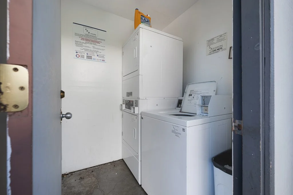 Small laundry room with stacked washer and dryer, a white laundry cart, and a trash can.