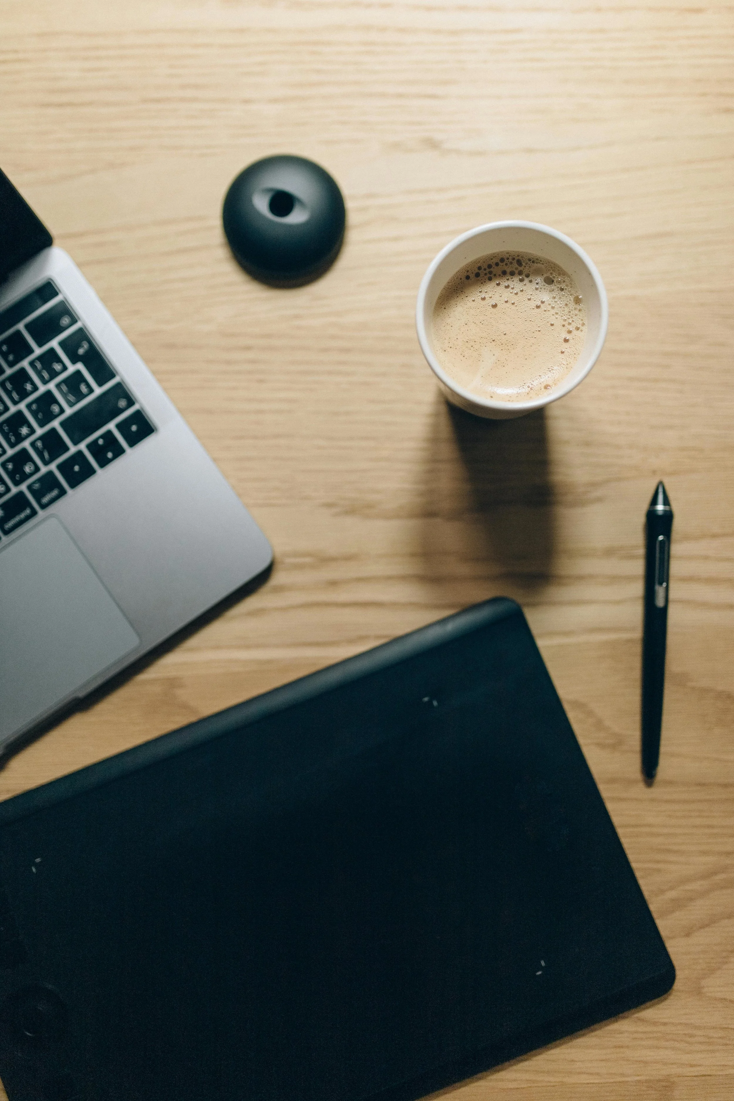 A desk with a laptop, black wireless speaker, disposable coffee cup, black notebook, pen, and a closed laptop. The surface is wooden.