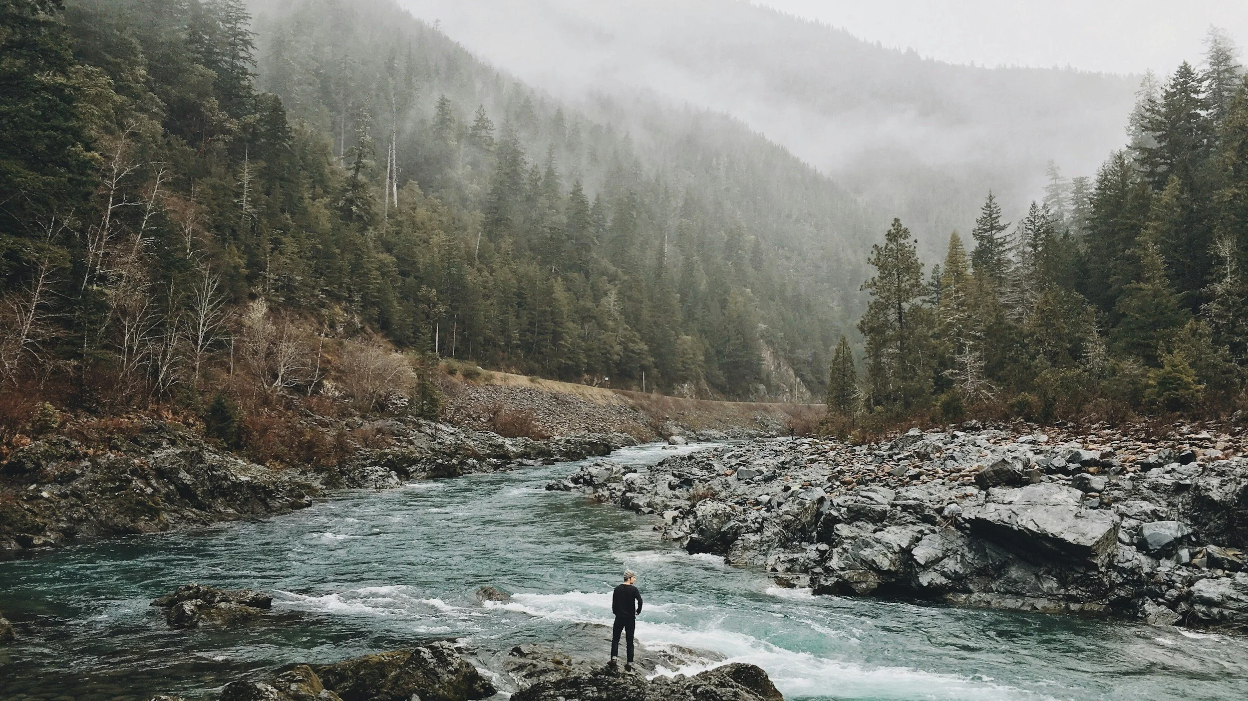 A person standing on rocks at the edge of a river in a forested mountainous area with misty peaks in the background.