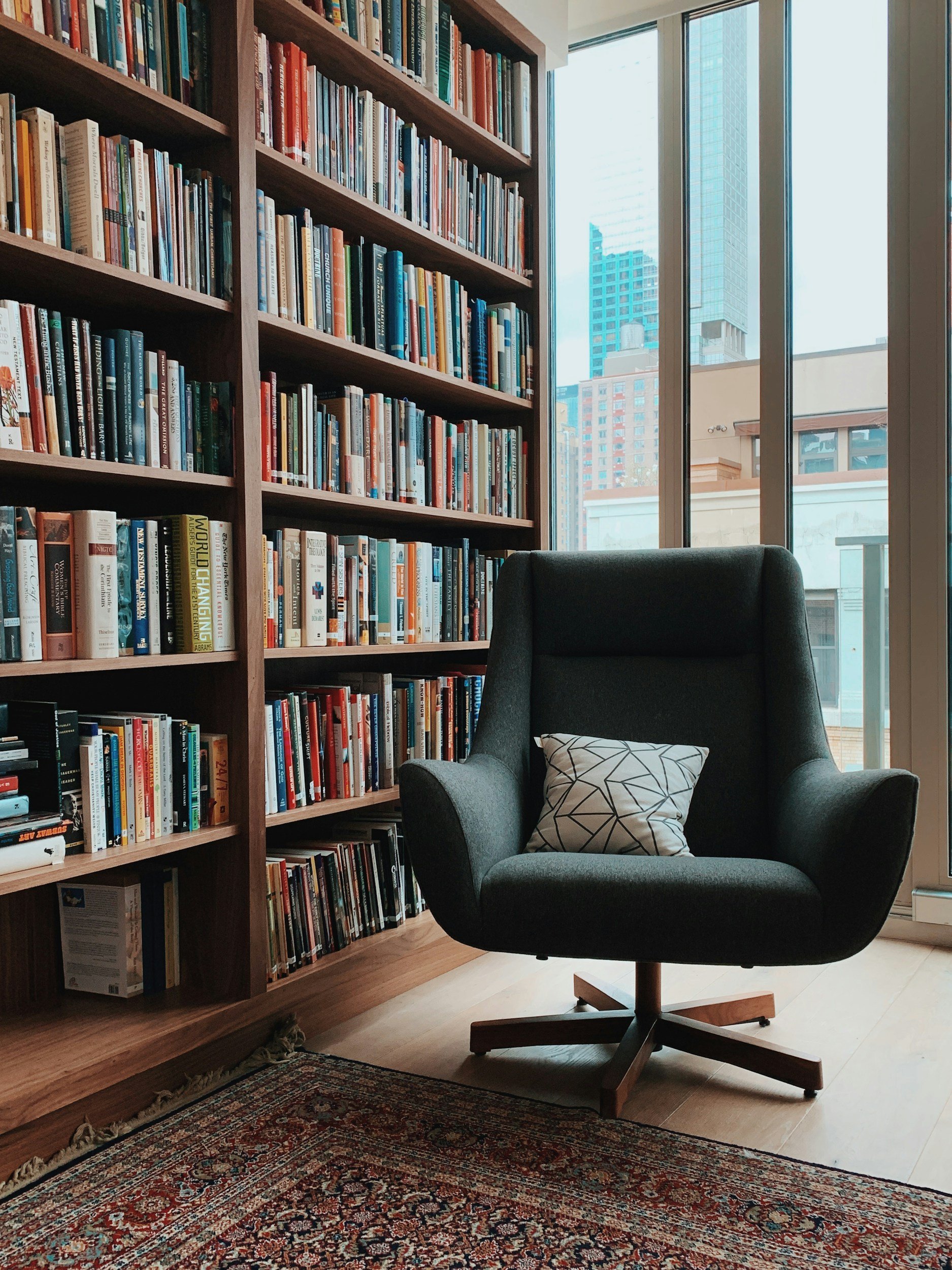 A cozy library corner with a tall wooden bookshelf filled with books on the left. A large window showing tall city buildings in the background is on the right. In front of the window is a black modern armchair with a patterned pillow on it, placed on a traditional Persian-style rug.