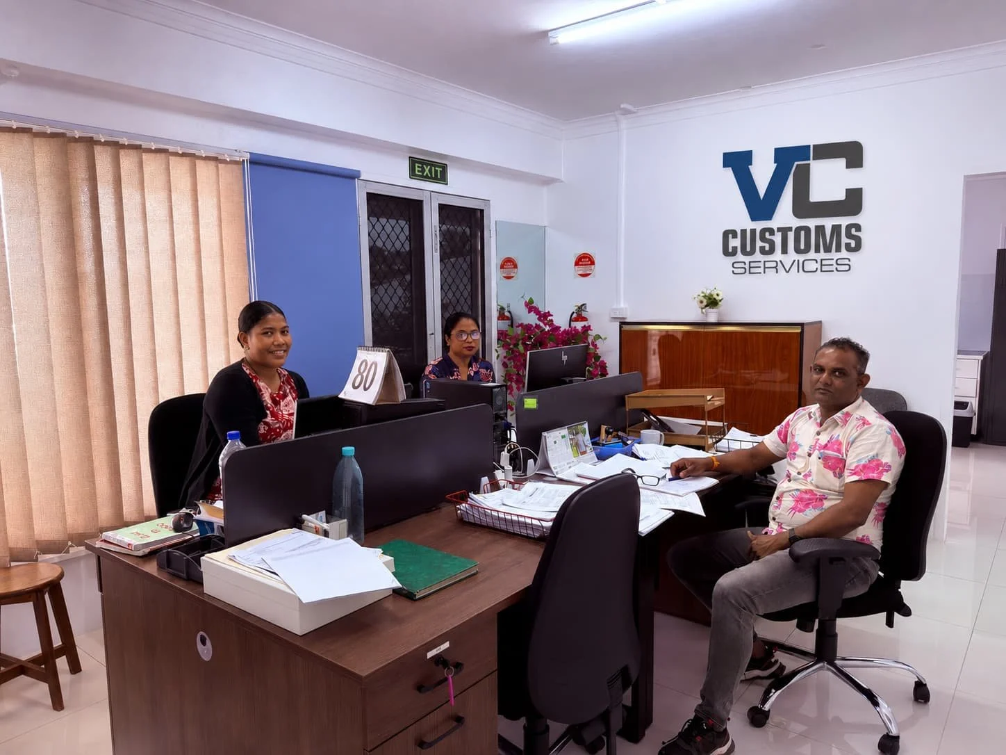 Office reception area with three employees sitting at desks. Two women on the left, one man on the right. The company logo 'VC Customs Services' on the wall behind them. Office supplies, papers, and water bottles on the desks, with curtains on the left side.