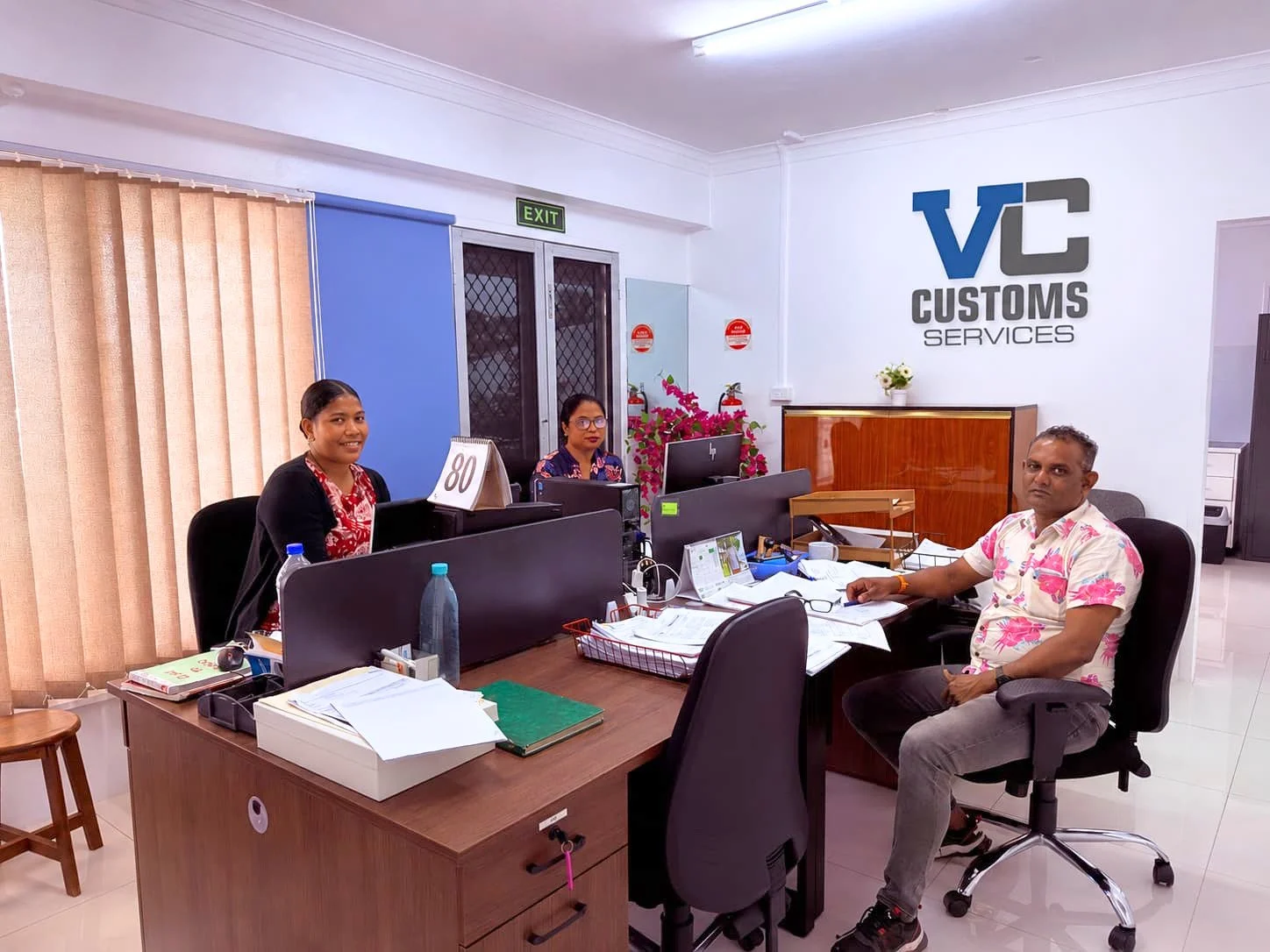 An office with three people sitting at desks inside VC Customs Services. Two women are behind the desks and one man is sitting in front of a desk with paperwork. There are computers, water bottles, and office supplies on the desks. The office has beige curtains, a wooden floor, and a white wall with