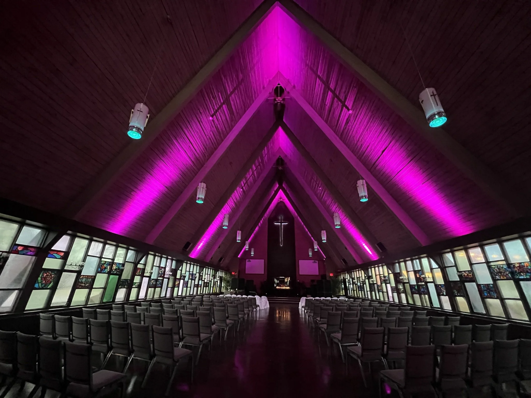 Church sanctuary interior with wooden ceiling, pink lighting accents, rows of black chairs, stained glass windows, and a cross at the front.