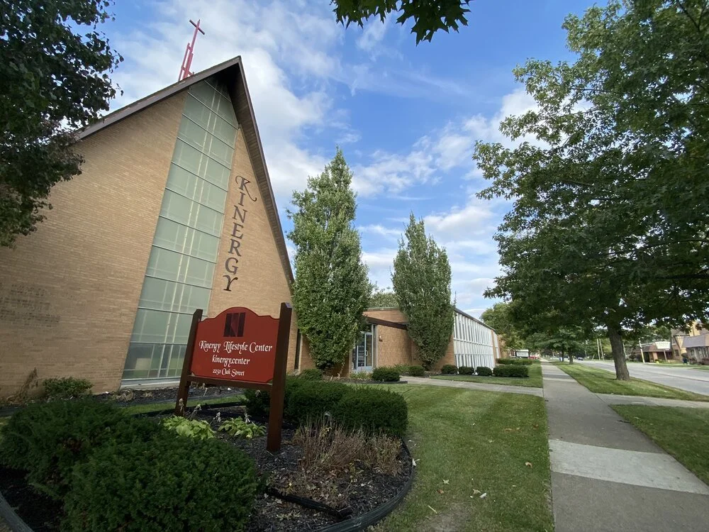 Exterior of a church building with a large glass window and a red cross on the roof. A red sign in front reads "Kunegry Lifestyle Center, 229 Oak Street." The church is surrounded by trees and a sidewalk, with a partly cloudy sky overhead.