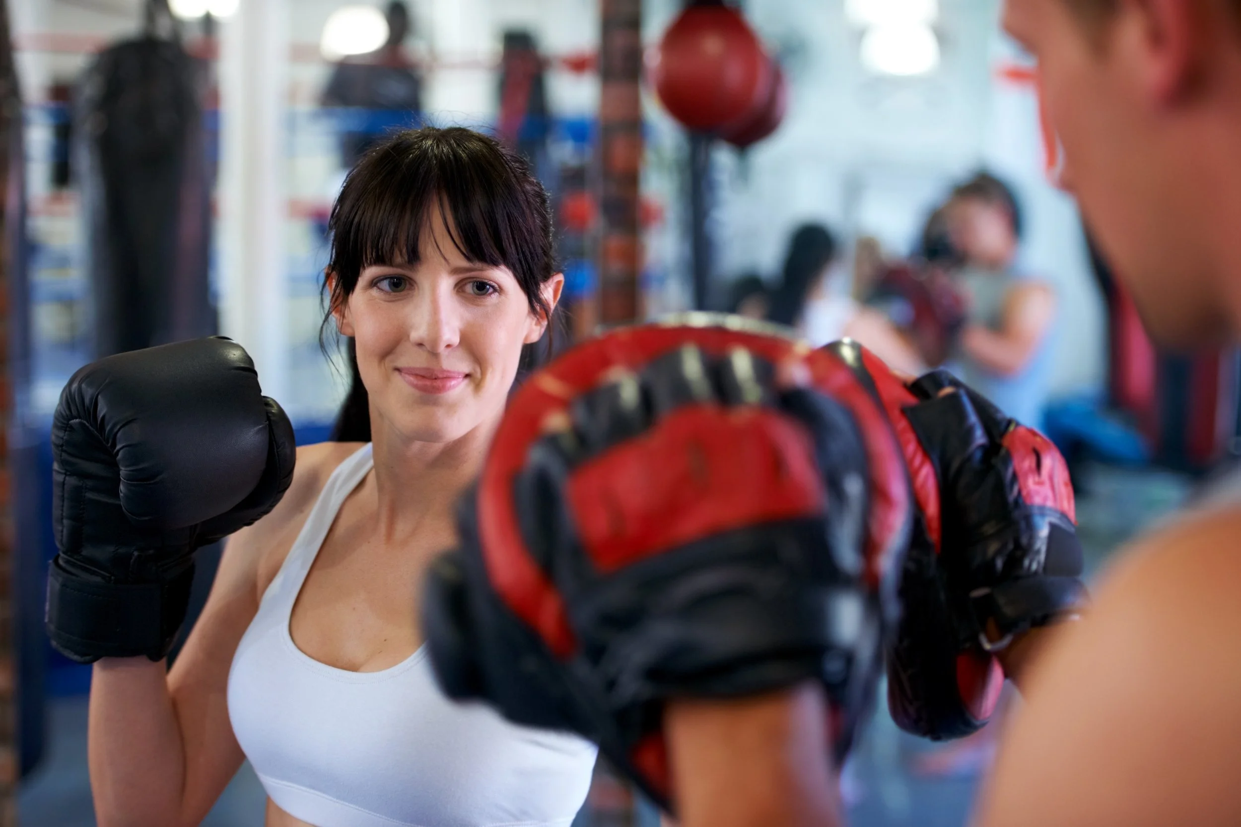 A woman with black hair and a white tank top is smiling while wearing boxing gloves and training in a gym. She is facing another person, possibly a trainer, who is holding focus mitts.