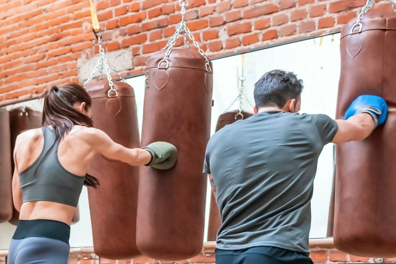 Two people boxing in a gym, practicing punches on brown punching bags hanging from a ceiling with a brick wall behind them.
