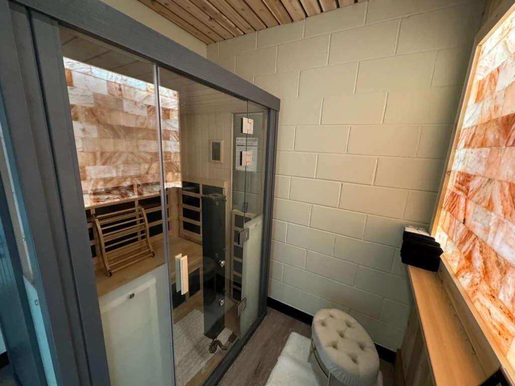 A small room with a glass-enclosed infrared sauna, a plush beige tufted ottoman, a wooden bench with black folded towels, a salt wall panel, and a white brick wall.