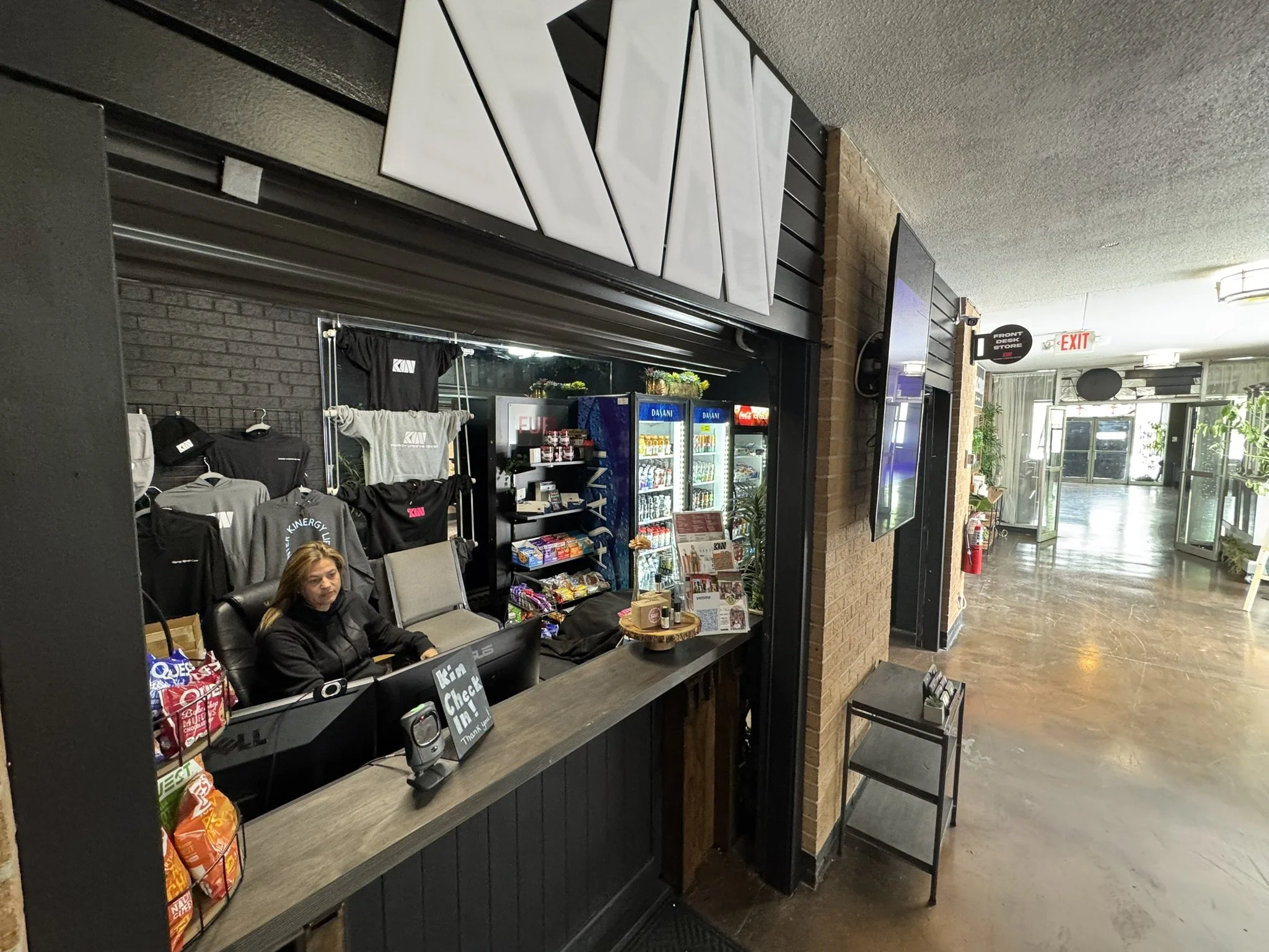 Inside a gift shop or checkout area featuring a woman seated behind the counter, merchandise on display including clothing and snacks, vending machines, and a sliding glass entrance door.