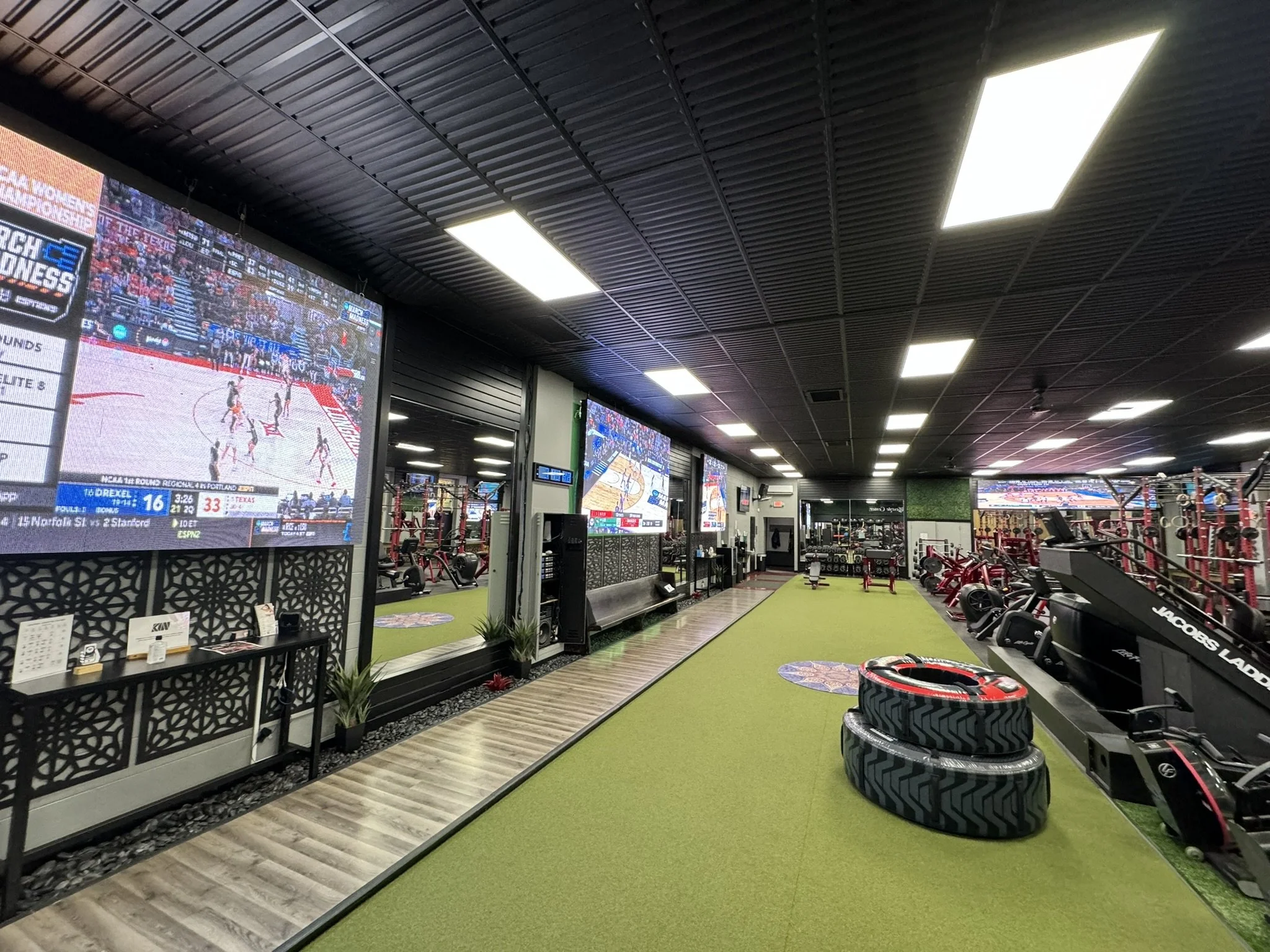 Inside a sports bar or lounge showing multiple large flat-screen TVs displaying a basketball game, with fitness or equipment area in the foreground featuring tires, weights, and exercise machines, and LED lighting on the ceiling.