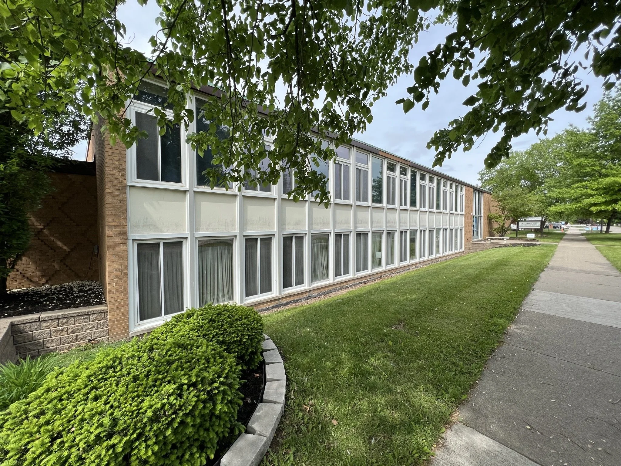 A sidewalk runs alongside a grassy area with hedges and trees in front of a brick building with large windows.